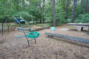 Spielplatz auf dem Campingplatz im Wald: grüne Rutsche, Sandkasten und Holzbank am Weg zwischen Bäumen.
