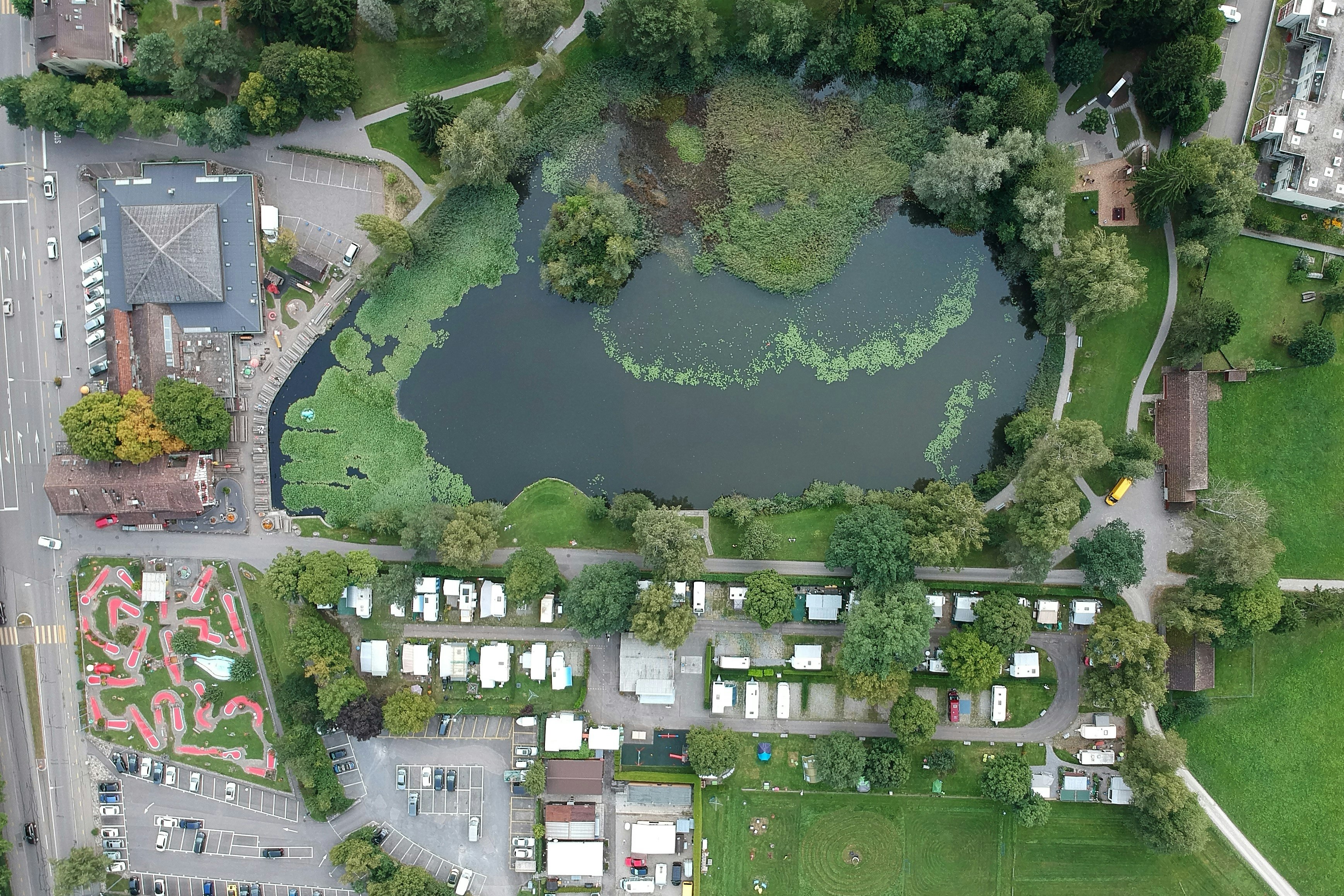 Luftaufnahme eines Campingplatzes mit See, Wegen und viel Grün, daneben Gebäude, Stellplätze und Wohnwagen.