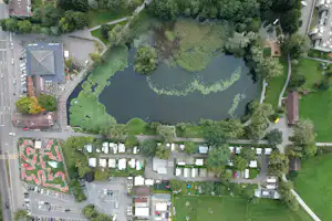 Luftaufnahme eines Campingplatzes mit See, Wegen und viel Grün, daneben Gebäude, Stellplätze und Wohnwagen.