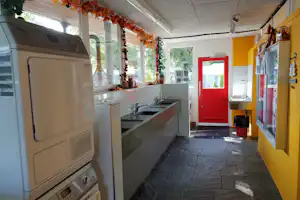 Camping-Küchenzeile mit Spüle, grauen Schränken und roten Eingangstür, große Fenster mit Blick ins Grüne.