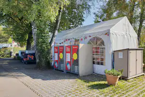 Großes Zelt auf dem Campingplatz zwischen Bäumen, mit Eingang und Absperrgittern sowie Schildern am Zaun.