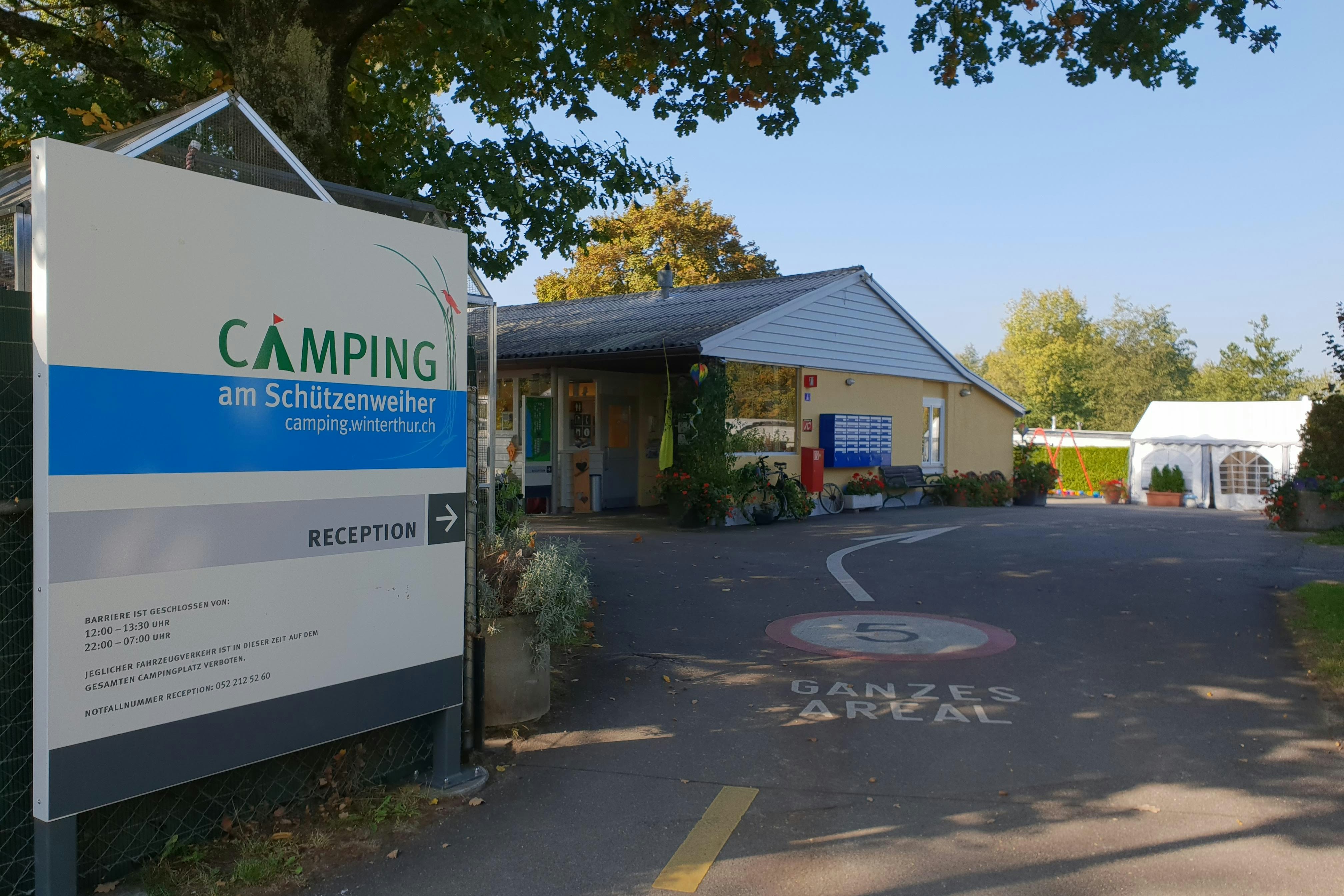 Schild „Camping am Schutzenweiher“ mit Pfeil zur Rezeption vor einem Gebäude und Parkplatz am Campingplatz.