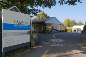 Schild „Camping am Schutzenweiher“ mit Pfeil zur Rezeption vor einem Gebäude und Parkplatz am Campingplatz.