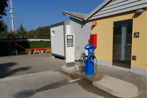 Camping-Anschlussstation mit blauer Pumpe, grünen Schläuchen und Beschilderung an einem Sanitärgebäude, daneben ein Chemie-Schild.