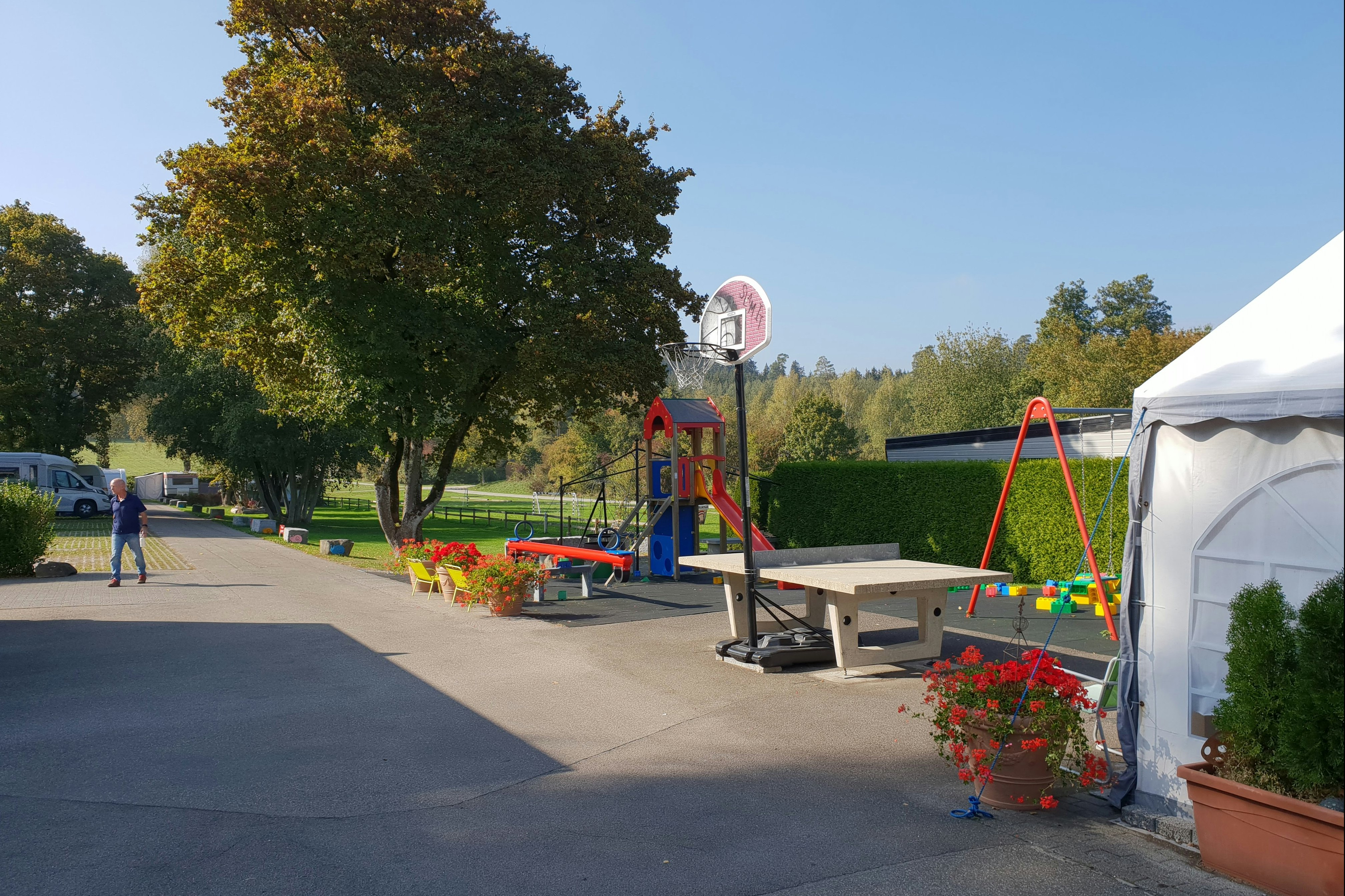 Spielplatz am Campingplatz mit Basketballkorb, Rutsche und Schaukel neben Wohnwagen und Zelt, umgeben von Hecken und Bäumen.