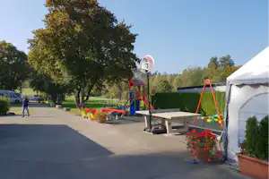 Spielplatz am Campingplatz mit Basketballkorb, Rutsche und Schaukel neben Wohnwagen und Zelt, umgeben von Hecken und Bäumen.