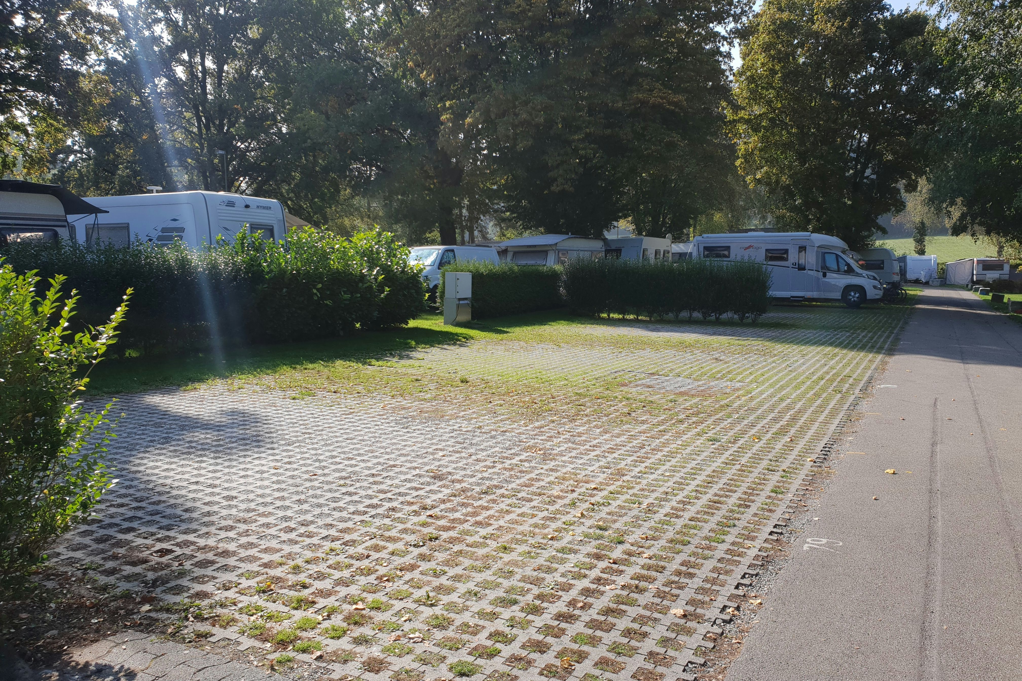 Wohnmobile und ein Wohnwagen stehen auf einem gepflasterten Stellplatz am Campingplatz, umgeben von Büschen und hohen Bäumen.