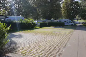 Wohnmobile und ein Wohnwagen stehen auf einem gepflasterten Stellplatz am Campingplatz, umgeben von Büschen und hohen Bäumen.