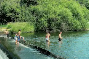 Fluss mit kleinem Wehr und Überlauf, mehrere Personen im Wasser am Campingplatz, umgeben von grünen Bäumen.