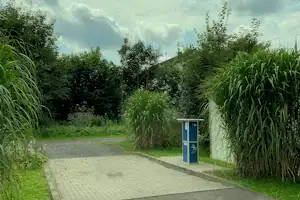 Gepflasterter Weg und Stellfläche am Campingplatz, umgeben von hohen Gräsern und Büschen, mit Infotafel am Rand.
