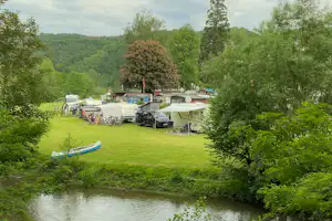 Campingplatz am Fluss mit Rasenfläche, mehreren Wohnmobilen und Mobilheimen, Bäumen und einem kleinen Steg am Ufer.