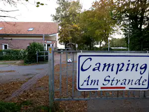Camping-Schild „Camping Am Strande“ am Eingang, daneben ein Zaun und ein Gebäude mit Ziegeldach und Bäumen im Hintergrund.