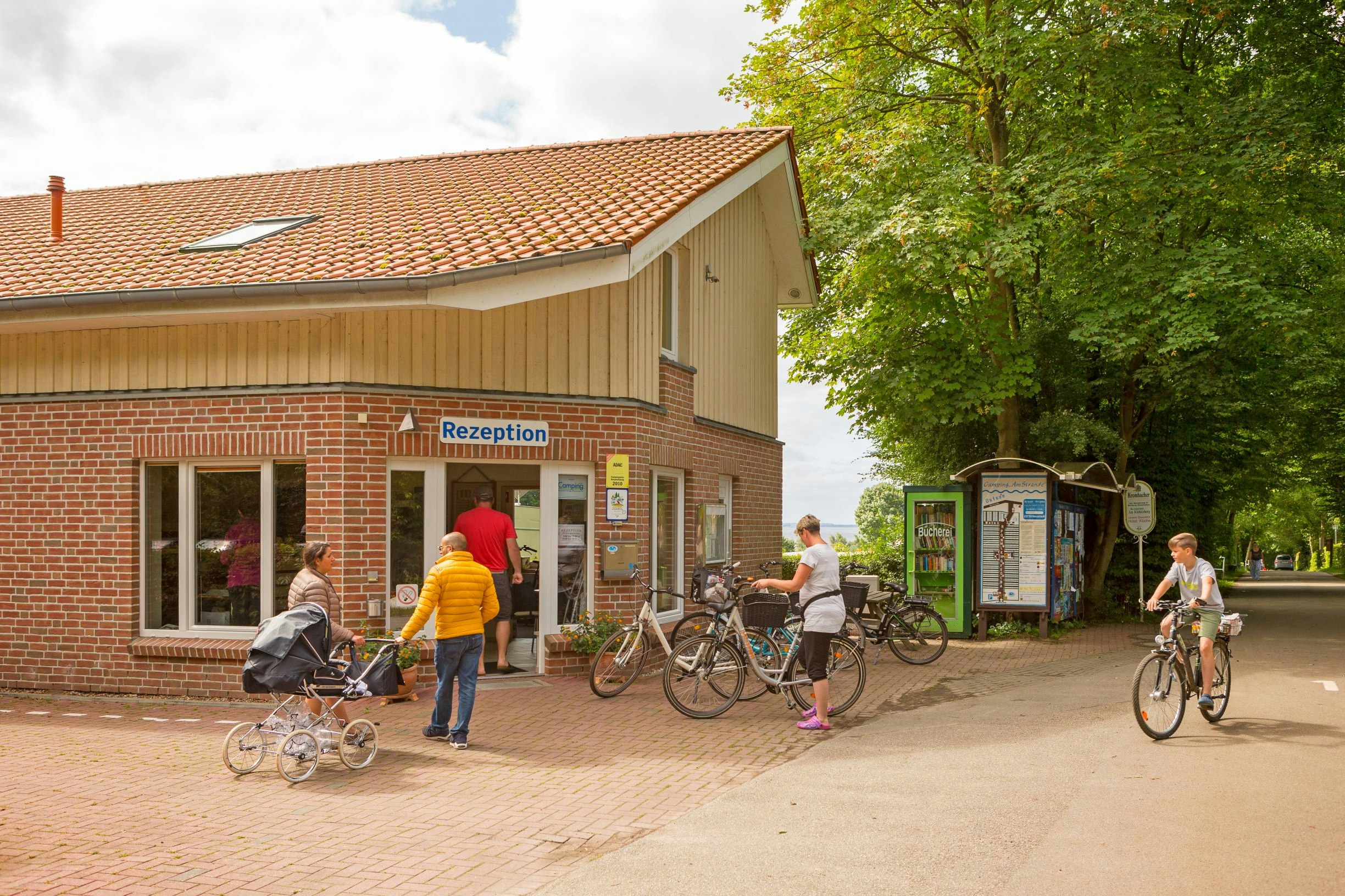 Rezeption eines Campingplatzes im Backsteingebäude, davor Fahrräder und Gäste auf dem Weg, Bäume im Hintergrund.
