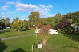 Stellplätze auf einer grünen Wiese mit Wohnwagen, Bäumen und einem Weg unter blauem Himmel.