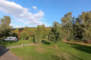 Grüner Campingplatz mit Rasenwegen, Bäumen und Hecken, im Hintergrund ein Wohnwagen und ein Wohnmobil am Stellplatz.