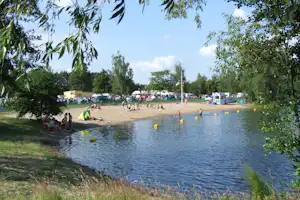 Badestrand am Campingplatz mit See, gelben Schwimmbojen, Zelten und Wohnmobilen hinter einem grünen Zaun.