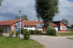 Campinggebäude mit roten Ziegeldächern, weißem Eingang und mehreren Fenstern, umgeben von Büschen, Rasen und einem Weg.