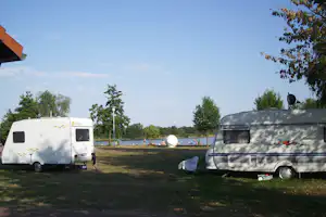 Zwei Wohnwagen auf einem Campingplatz am See, Rasenfläche, Bäume und ein Wohnmobil im Vordergrund.