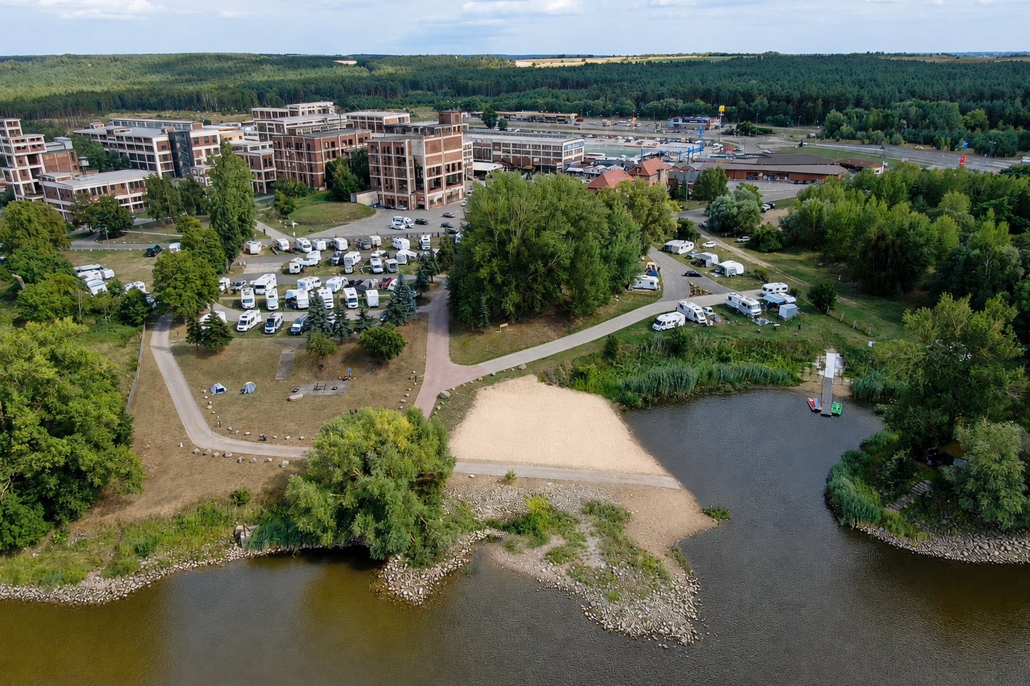 Luftaufnahme eines Campingplatzes mit Wohngebäuden, Stellplätzen, Wegen und einem kleinen See im Vordergrund.