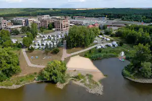 Luftaufnahme eines Campingplatzes mit Wohngebäuden, Stellplätzen, Wegen und einem kleinen See im Vordergrund.