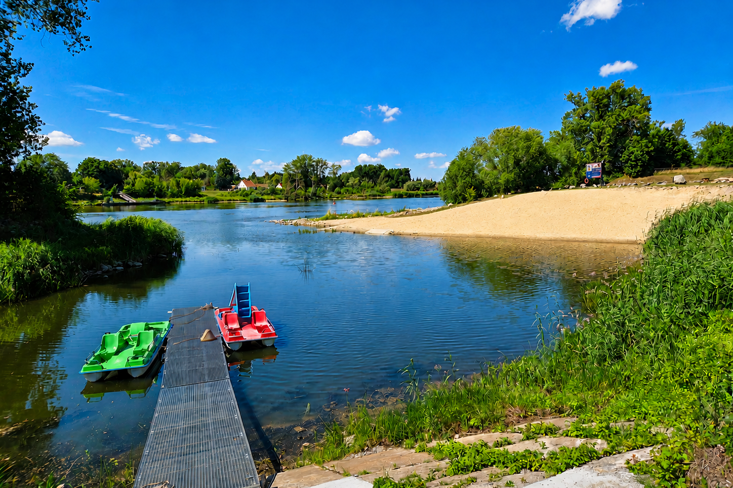 Bootssteg am See mit zwei Booten, grünen Ufern und einem Sandstrand, im Hintergrund Bäume und ein Gebäude.