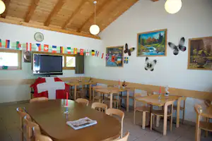 Innenraum mit Holzbalkendecke, Tischen und Stühlen, TV-Gerät und vielen Länderflaggen an der Wand.