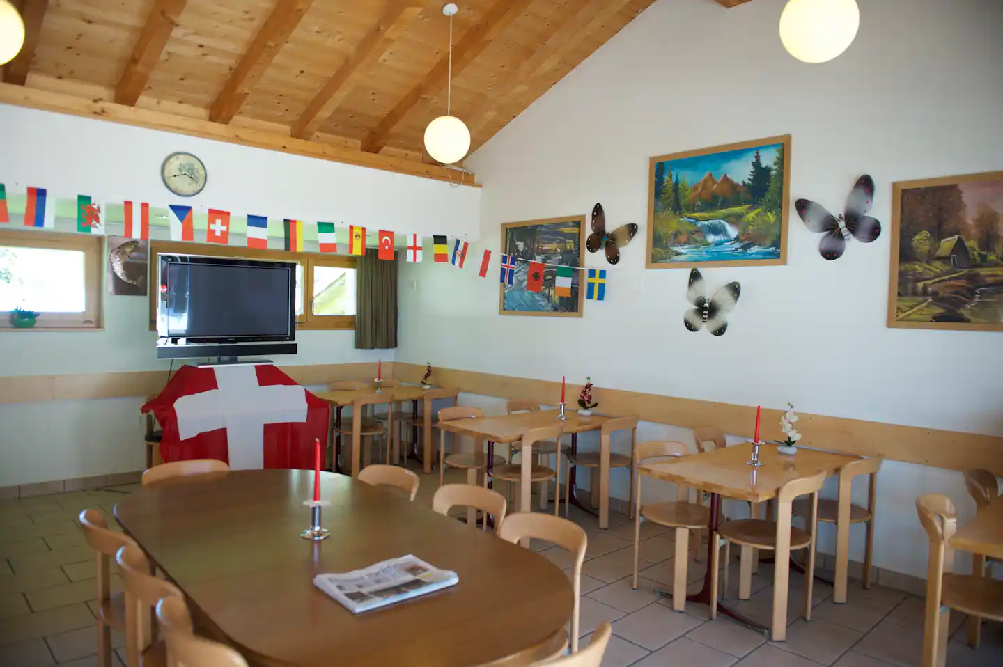 Innenraum mit Holzbalkendecke, Tischen und Stühlen, TV-Gerät und vielen Länderflaggen an der Wand.