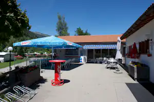 Terrasse am Campingplatz mit blauen Sonnenschirmen, Stühlen und Tischen vor einem Gebäude, im Hintergrund Berge und Bäume.