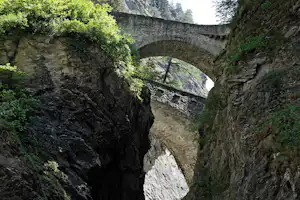 Steinbrücke mit Bogen über einem engen Felsental, Kopfsteinpflasterweg und Geländer, umgeben von Bäumen und Felswänden.