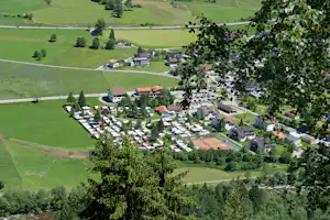 Luftaufnahme einer Campinganlage mit Wohngebäuden, Stellplätzen und Parkplatz in grünen Hügeln, umgeben von Wiesen und Bäumen.
