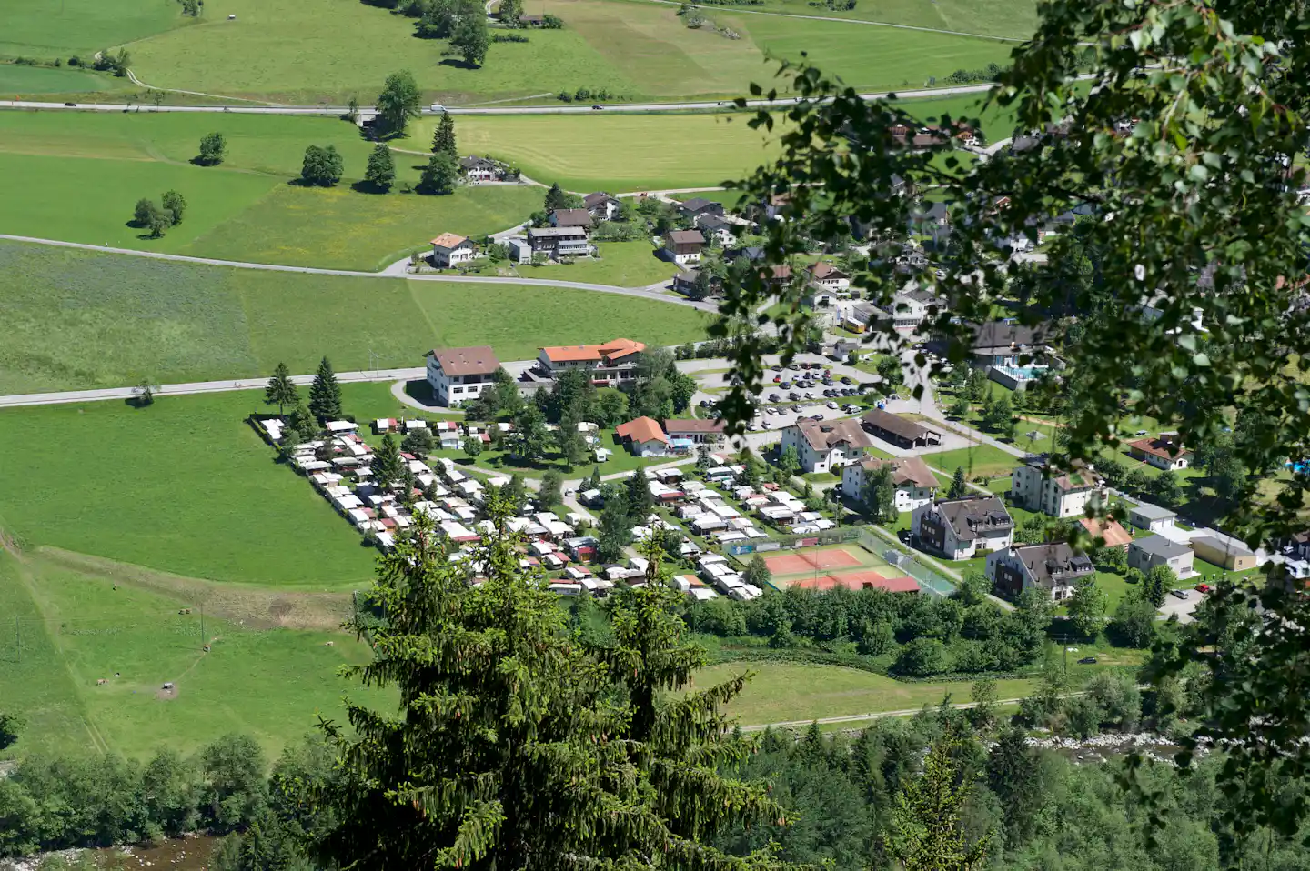 Luftaufnahme einer Campinganlage mit Wohngebäuden, Stellplätzen und Parkplatz in grünen Hügeln, umgeben von Wiesen und Bäumen.