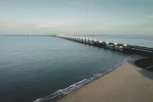 Langer Steg im Hafen über dem Wasser, mit Uferpromenade und Windrädern in der Ferne.