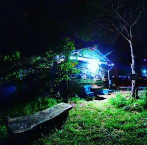 Campingplatz bei Nacht: beleuchtetes Gebäude mit Holzveranda, Tischen und Stühlen im Garten, umgeben von Bäumen.
