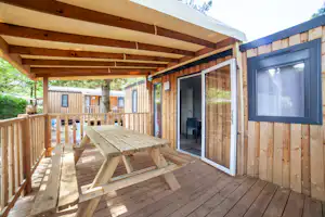 Holzterrasse mit Tisch und Stühlen, überdachtem Bereich und offenen Türen am Holzbungalow, im Hintergrund Bäume.