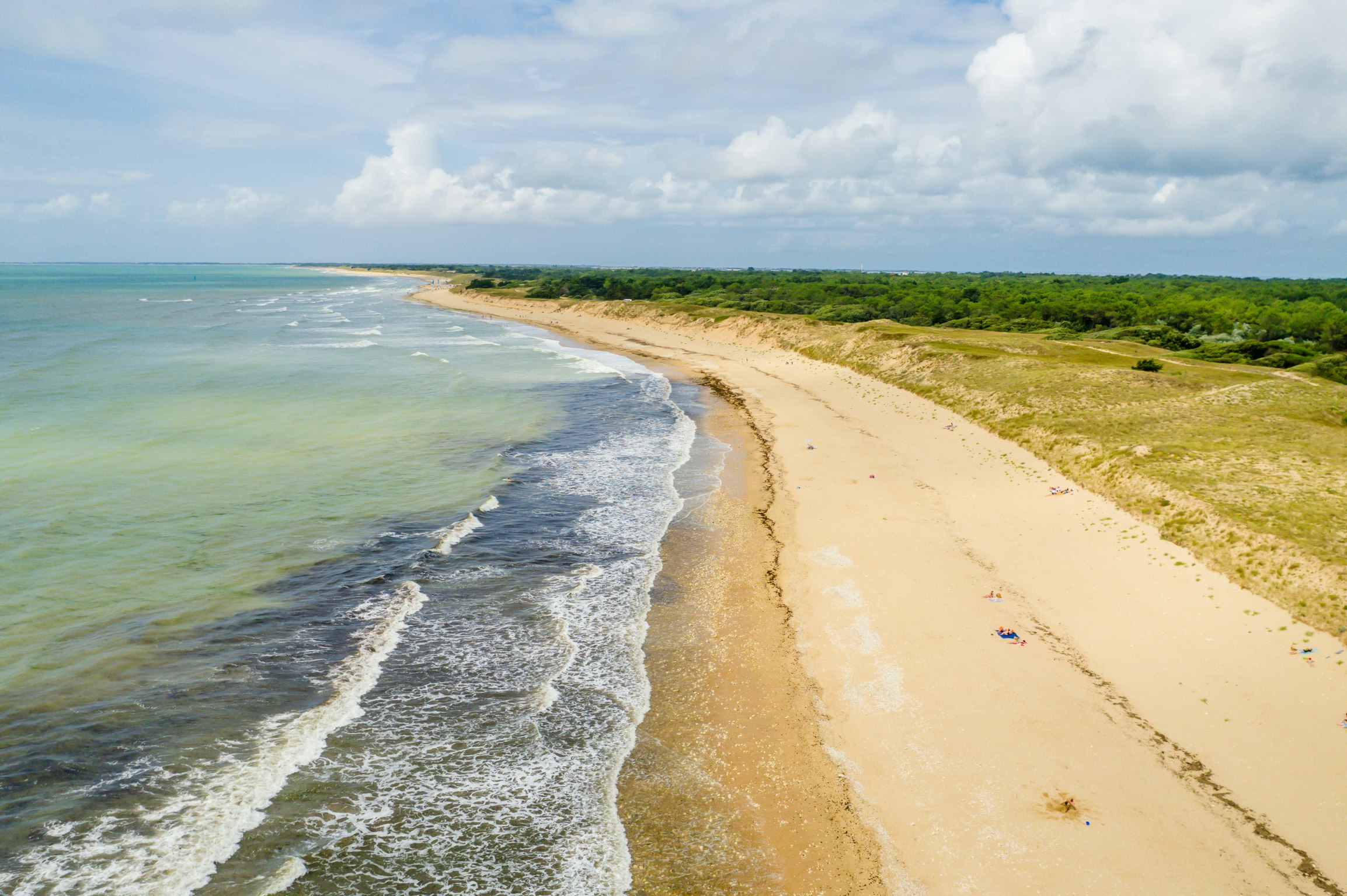 Luftaufnahme von einem langen Sandstrand an der Küste mit Wellen im Meer und grünen Dünen im Hintergrund.