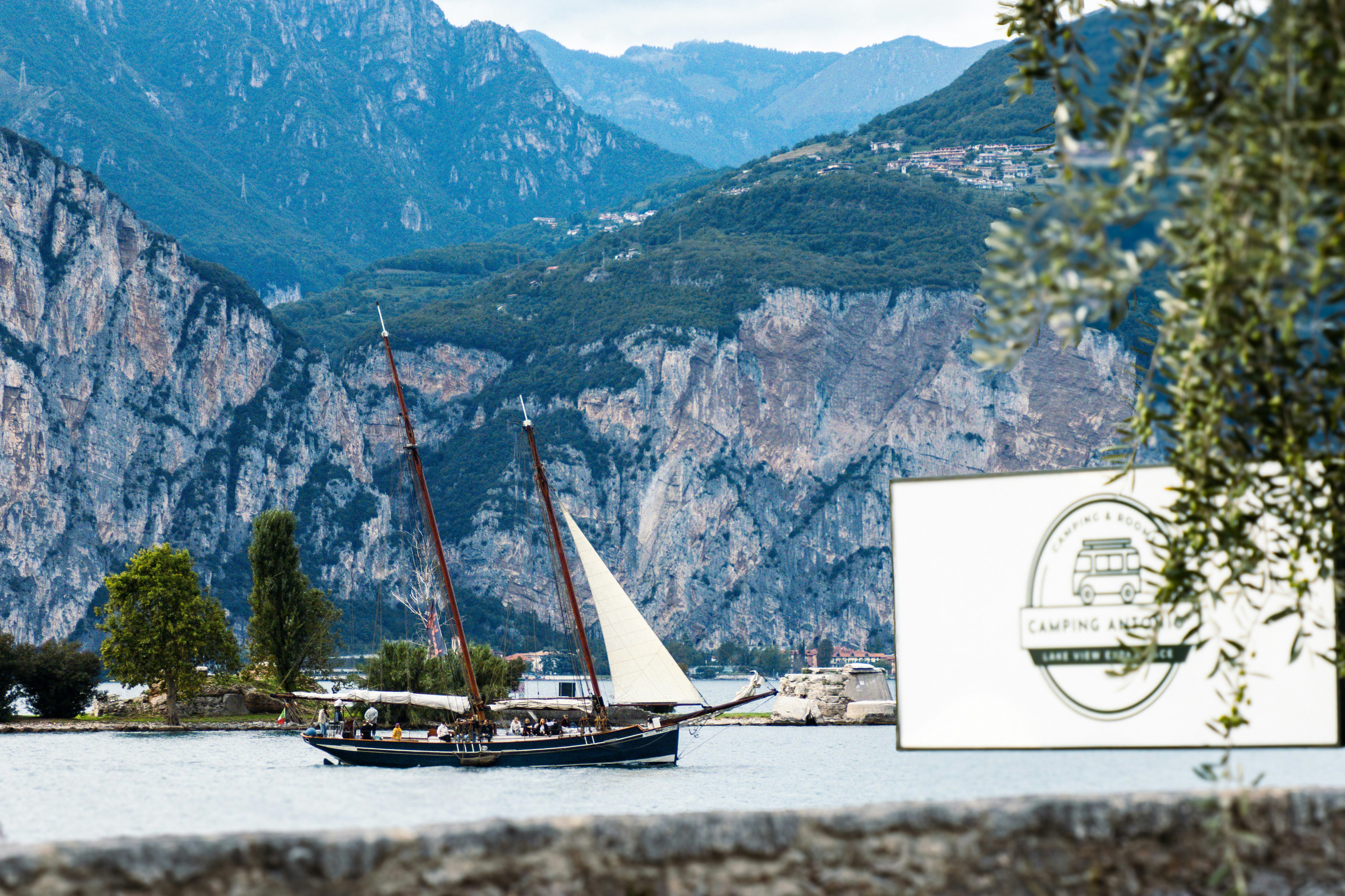 Segelboote am See vor einer Bergkulisse, im Vordergrund ein Schild am Ufer mit Bäumen und Steinstützmauer.