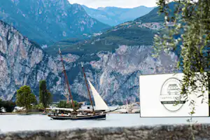 Segelboote am See vor einer Bergkulisse, im Vordergrund ein Schild am Ufer mit Bäumen und Steinstützmauer.