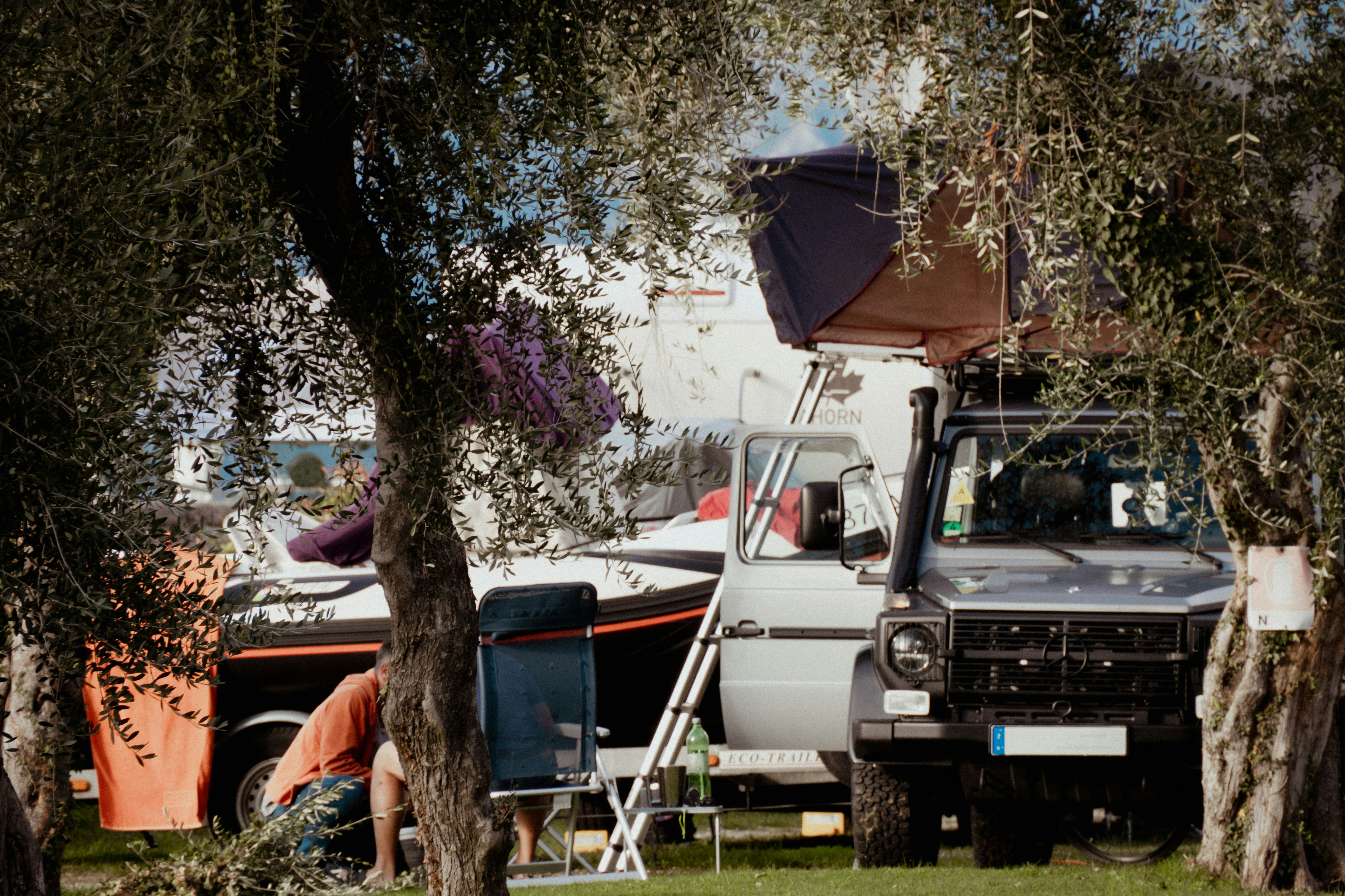 Wohnmobil auf einem Campingplatz zwischen Bäumen, mit Markise, Leiter und Campingstühlen am Stellplatz.