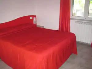 Bett mit roter Bettwäsche im hellen Zimmer, roter Vorhang und Fenster mit Blick auf den Außenbereich.