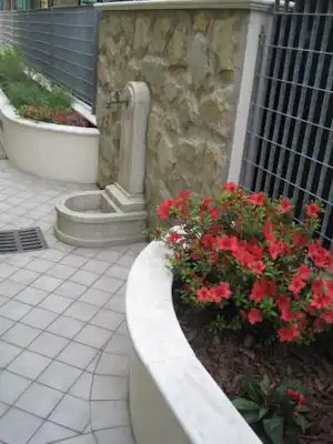 Steinwand mit Brunnen und Blumenbeet auf einer gepflasterten Terrasse am Campingplatz, neben Metallgeländer und Pflanzkübeln.