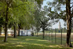 Mobilheime auf dem Campingplatz zwischen hohen Bäumen, mit Gartenrasen, Zaun und Blick auf einen Kirchturm in der Ferne.
