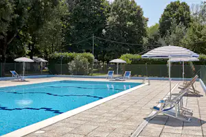 Camping-Pool mit blauem Wasser, gepflasterter Terrasse und mehreren Liegen unter Sonnenschirmen im Grünen.