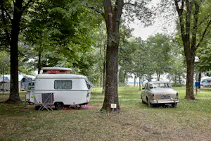 Wohnwagen und Auto auf einem Campingplatz zwischen Bäumen, mit Vorzelt und Stellplatzschild im Gras.