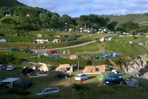 Campingplatz auf einer grünen Wiese mit vielen Zelten und geparkten Autos, umgeben von Bäumen und Hügeln.
