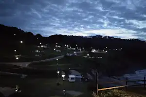 Campingplatz bei Abendlicht: beleuchtete Stellplätze mit Wohnwagen und Zelten, Weg und Hecken vor dunklen Hügeln.