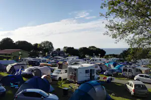 Campingplatz mit vielen Zelten und Wohnmobilen, dahinter Bäume und das Meer am Horizont unter blauem Himmel.