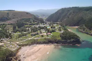 Luftaufnahme eines Campingplatzes am Strand in einer grünen Talsenke mit vielen Stellplätzen, Autos und türkisfarbenem Meer.