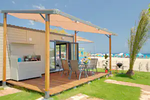 Wohnzelt mit Holzterrasse, Markise und Sitzgruppe am Campingplatz, im Hintergrund Meer und Palmen.