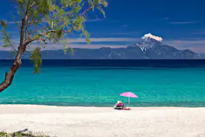 Türkisfarbenes Meer und Sandstrand mit rosa Sonnenschirm, davor ein kleines Boot und im Hintergrund Berge.