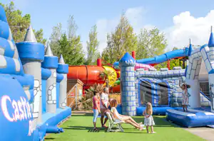Blaues Hüpfburg-Spielgerät auf dem Campingplatz mit Rasen, Kindern und Erwachsenen im Hintergrund.